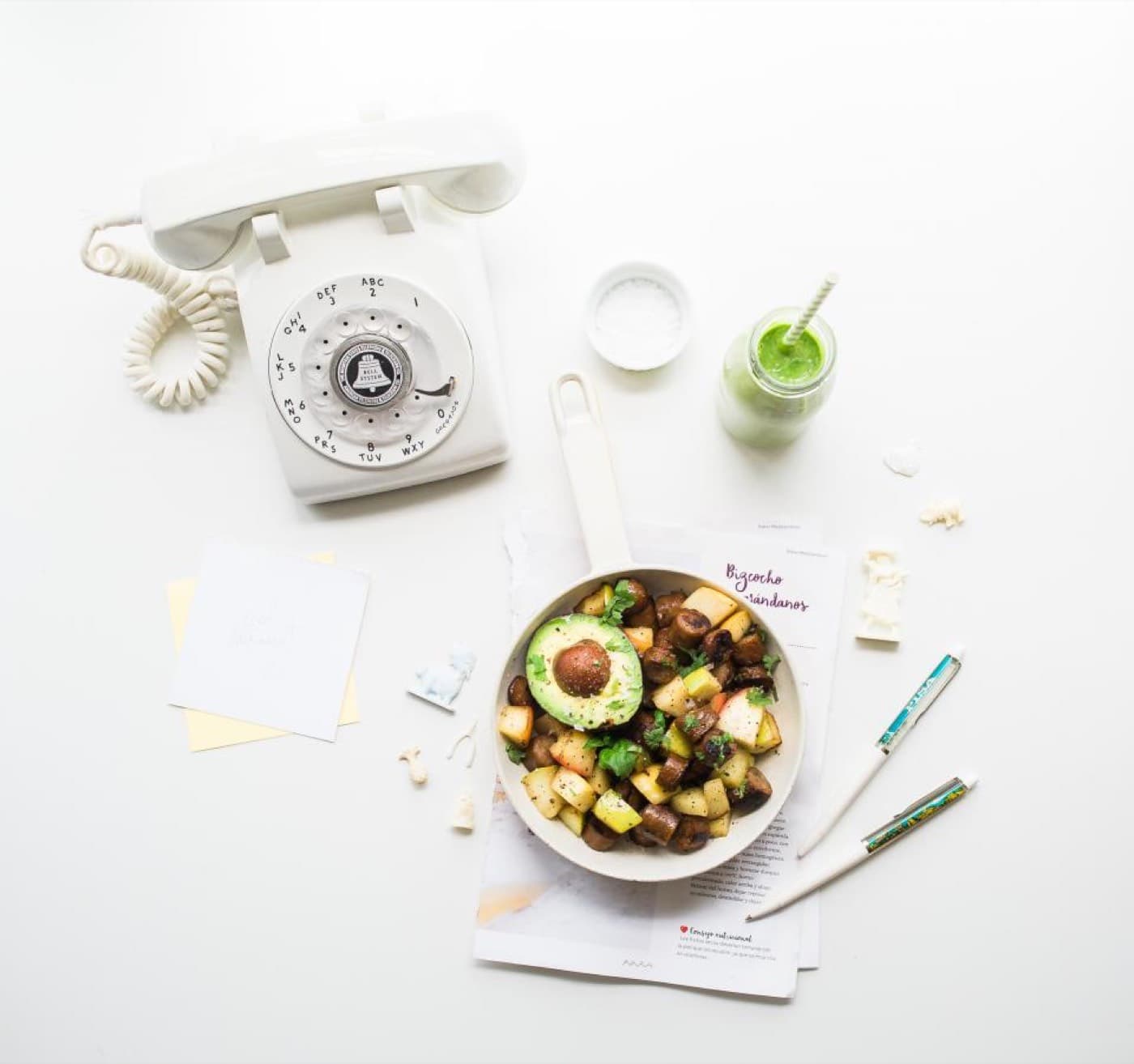 Vue de dessus d'un verre de smoothie vert posé près d'un bol d'avocat et de légumes rôtis, sur une table blanche